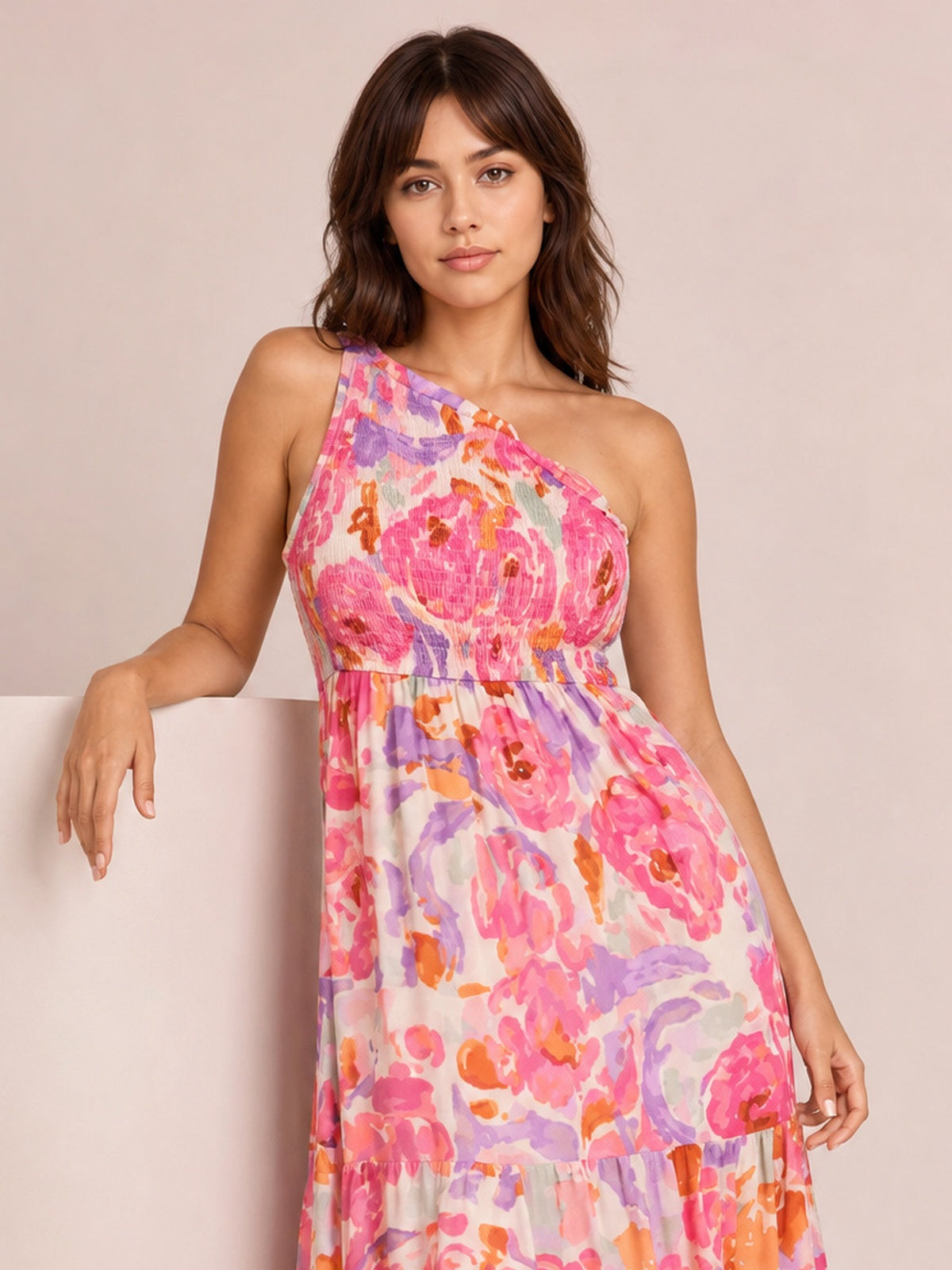 A model poses in a vibrant floral print maxi dress with a one-shoulder neckline, smocked bodice, and flowing tiered silhouette in pink, coral, orange, and lavender.