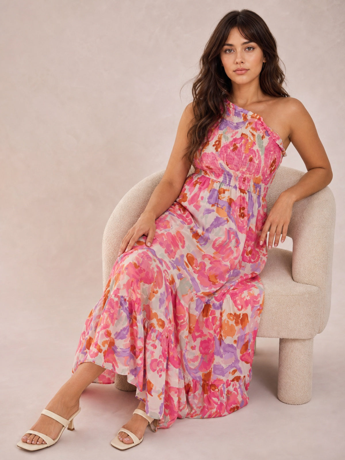 Model sits with one hand on her lap in a vibrant floral print one-shoulder maxi dress featuring a shirred bodice and flowing tiered skirt.