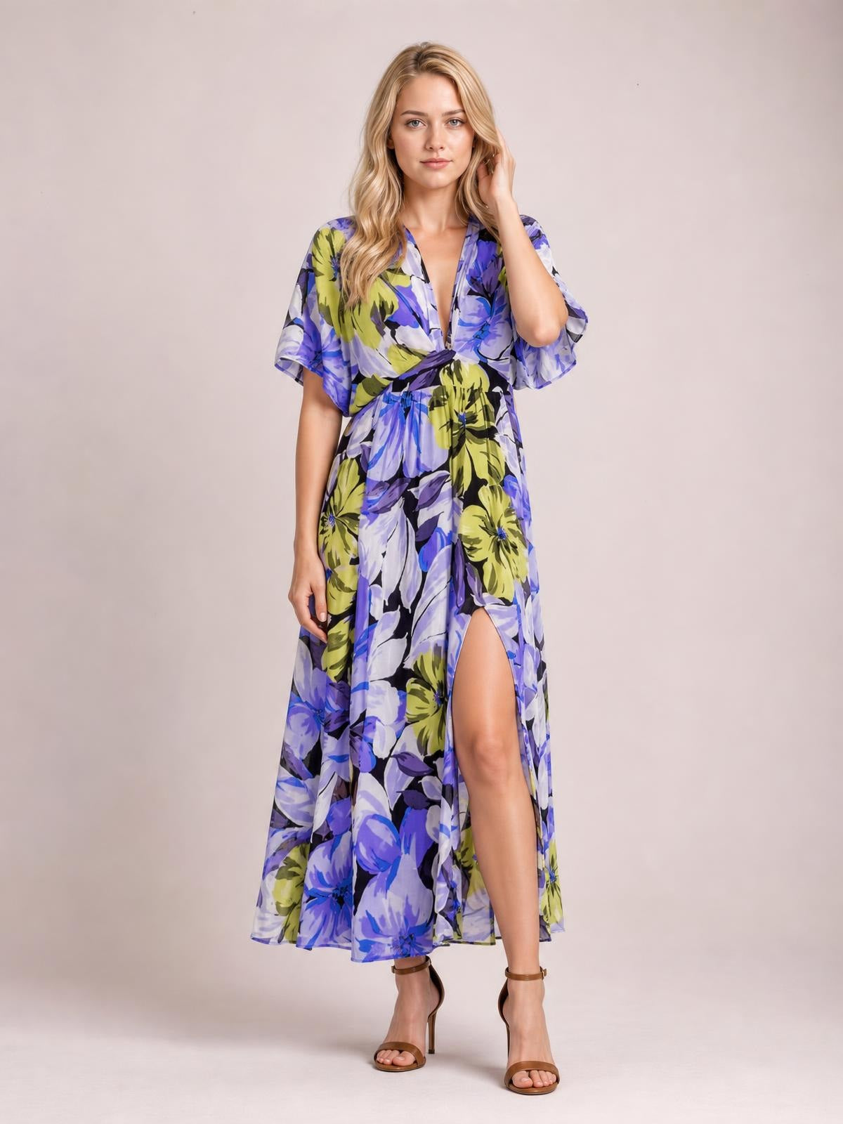Model poses in a blue, purple, and lime floral print V-neck maxi dress with flutter sleeves, a twisted waist, flowing silhouette, and high front slit.