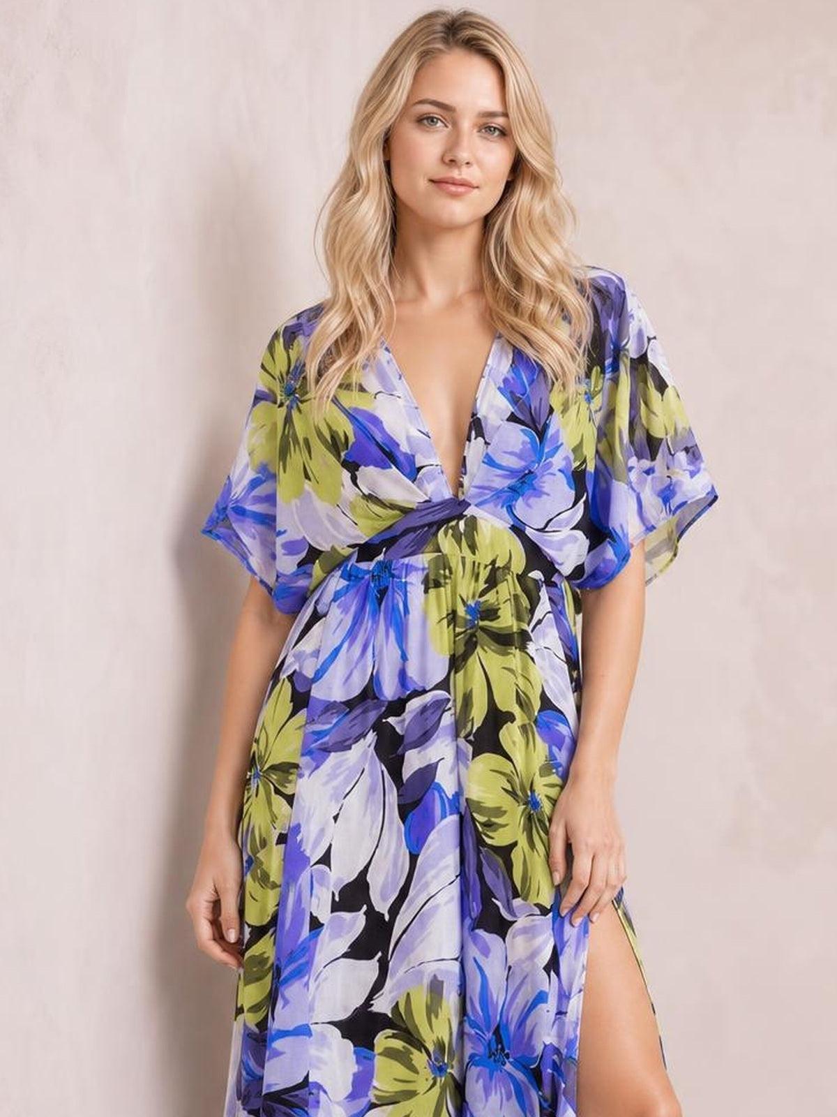 A model stands in a flowing floral print maxi dress with a deep V-neck, twisted waist, loose short sleeves, blue, purple and lime hues, and a high slit.