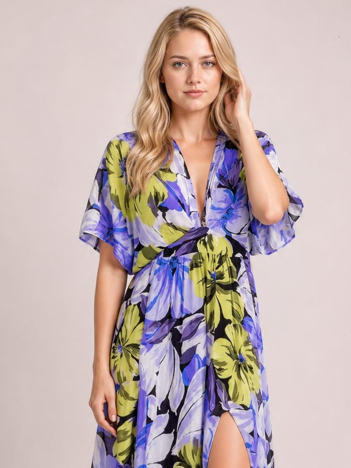 A model poses in a colorful floral print V-neck maxi dress with a twisted waist, flutter sleeves, flowing silhouette, and high slit revealing one leg.