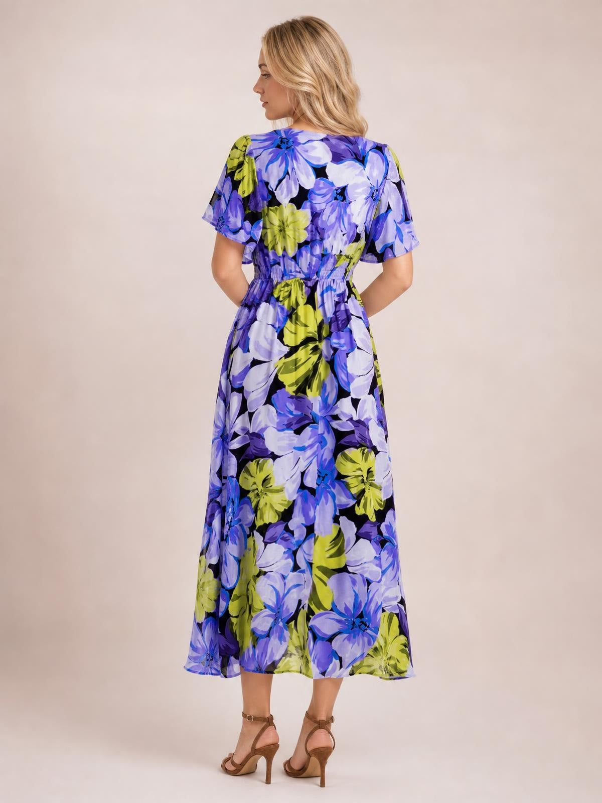 Back view of a model in a blue, purple, and lime floral print maxi dress with short sleeves, a smocked waist, and a flowing A-line silhouette.