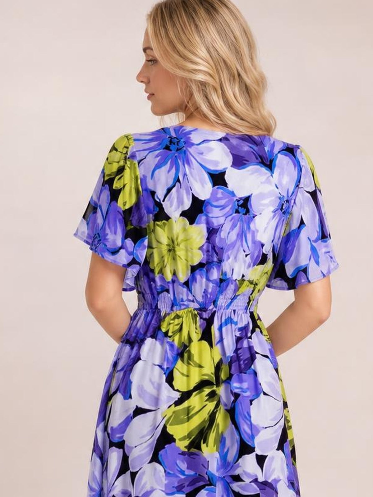 Model poses with hands at her waist, showcasing the back of a blue, purple, white, and lime floral print maxi dress with short sleeves and a gathered waist.