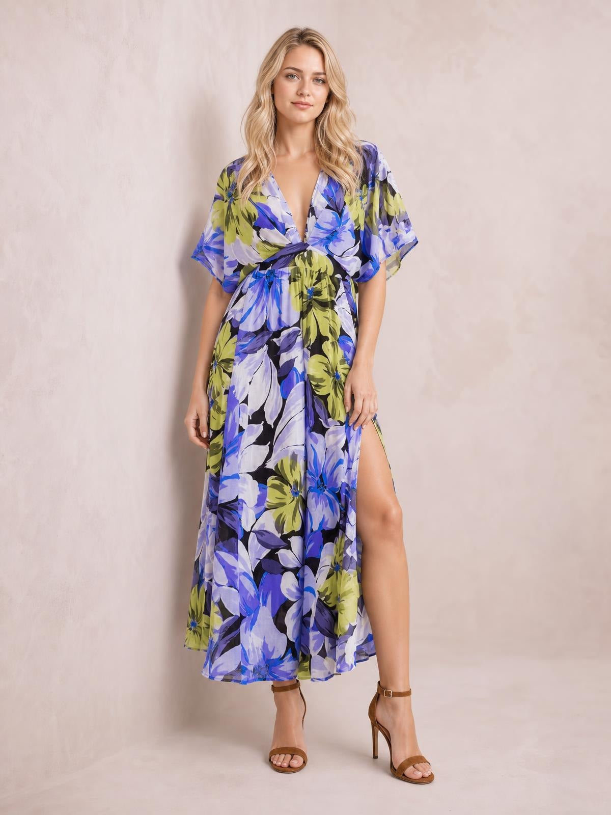 Model wearing a blue, purple, and olive floral print maxi dress with a deep V-neck, twisted waist, flutter sleeves, flowing silhouette, and high side slit.