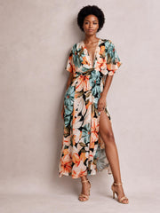 Model poses in a floral print V-neck maxi dress with a twisted waist, short sleeves, flowing silhouette, and high side slit in teal, coral, cream, and black.