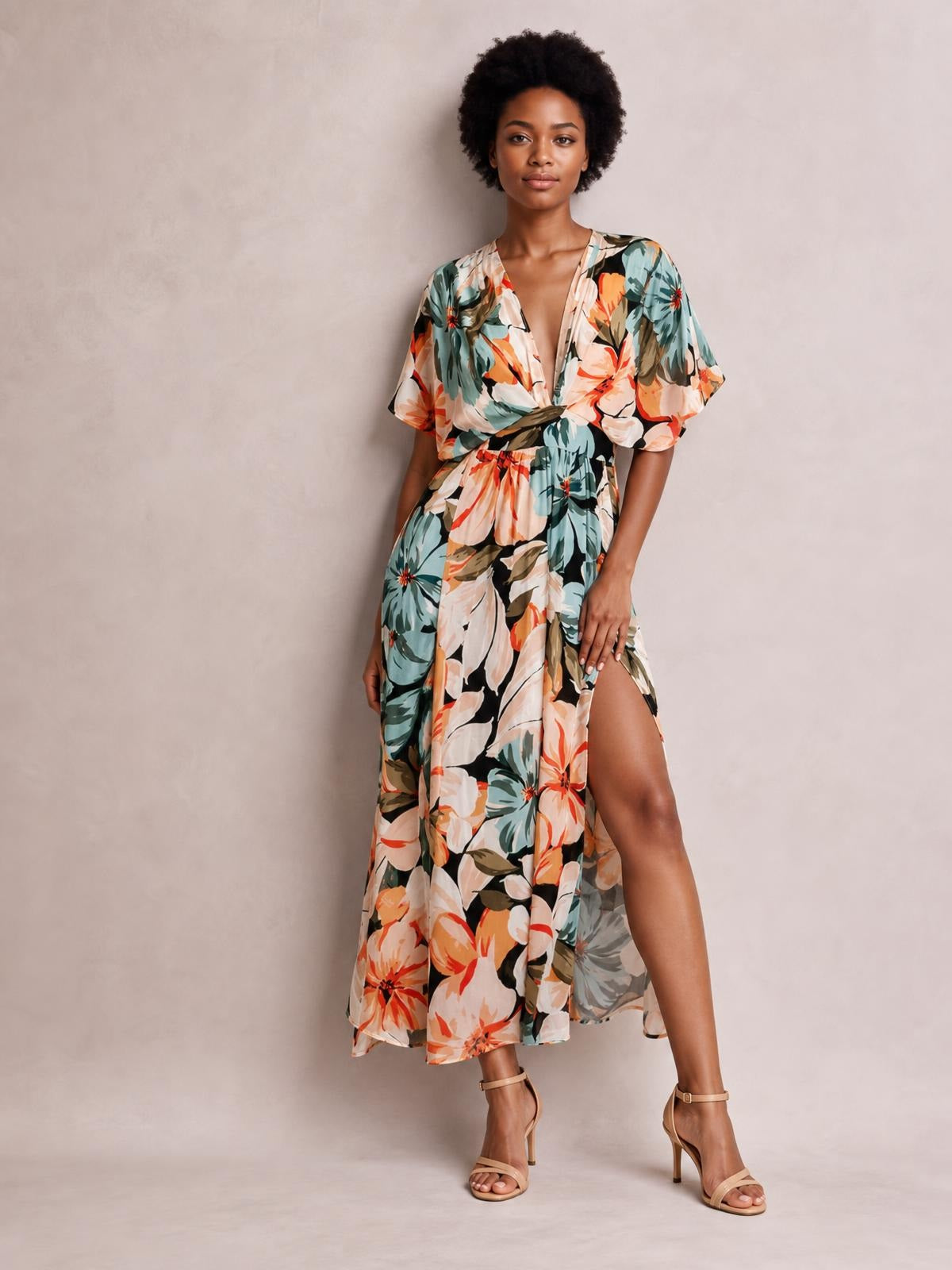 Model poses in a floral print V-neck maxi dress with a twisted waist, short sleeves, flowing silhouette, and high side slit in teal, coral, cream, and black.