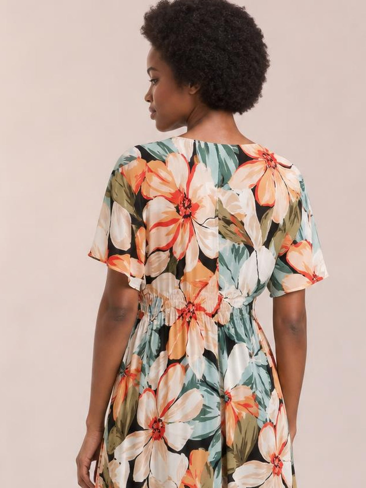 A model poses in a colorful floral-print maxi dress with short sleeves, a gathered waist, and a flowing silhouette, shown from the back.