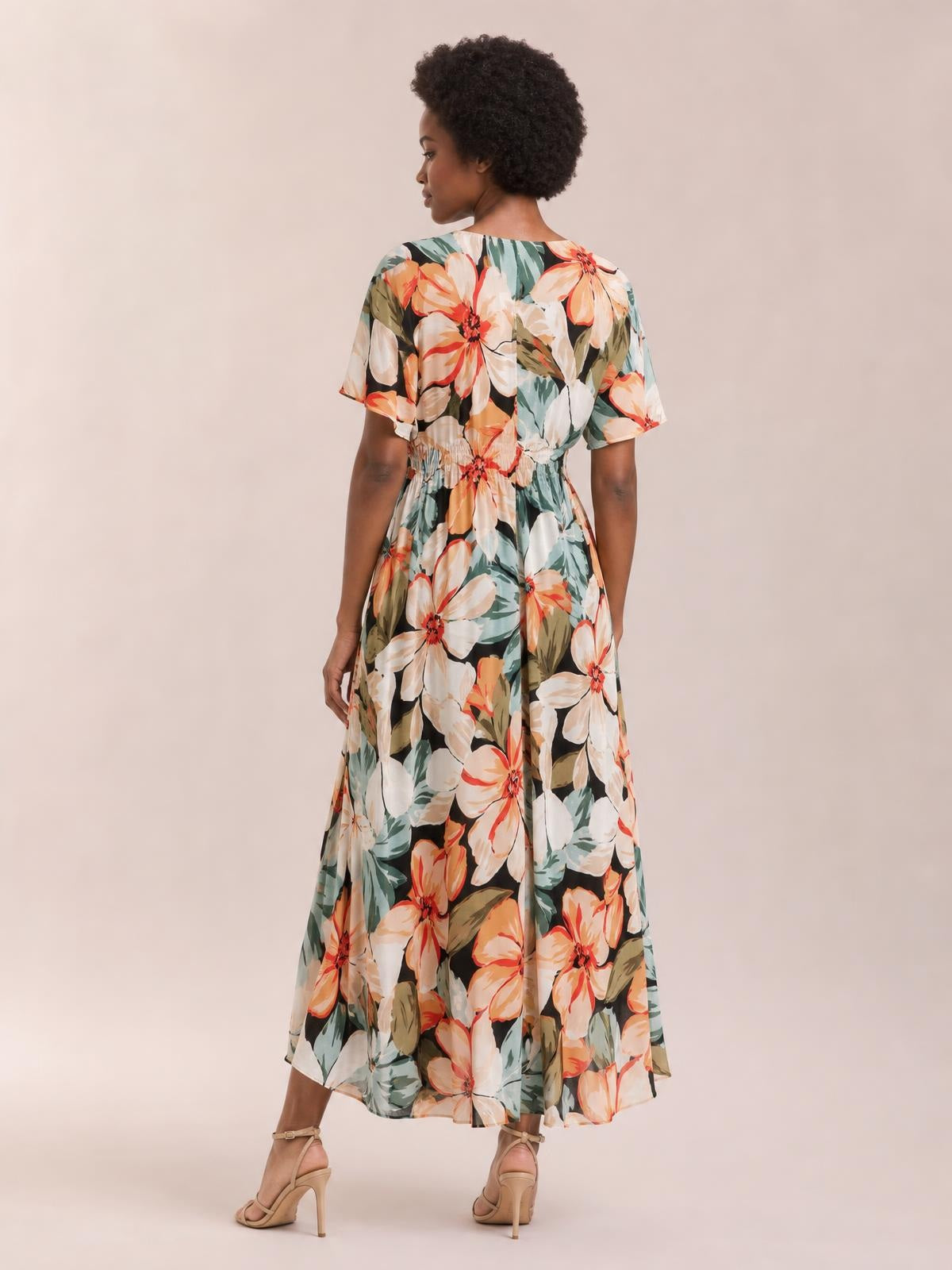 A model looks over her shoulder in a multicolor floral print maxi dress with short sleeves, a smocked waist, and a flowing ankle-length silhouette.