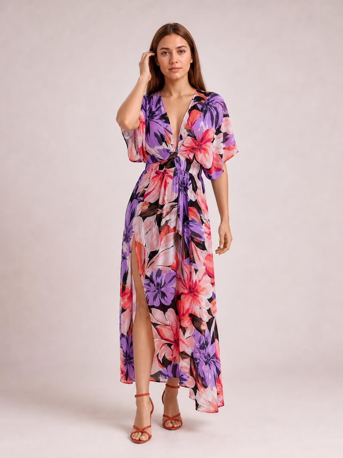 A model stands touching her hair in a vibrant purple, pink, and black floral print maxi dress with a V-neck, twisted waist, flutter sleeves, and high slit.