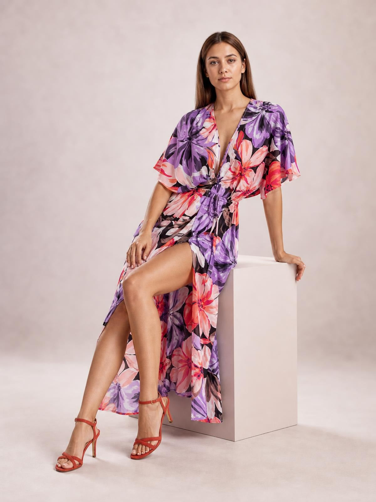 A seated model wears a purple and pink floral print V-neck maxi dress with flutter sleeves, a twisted waist, and a high slit.