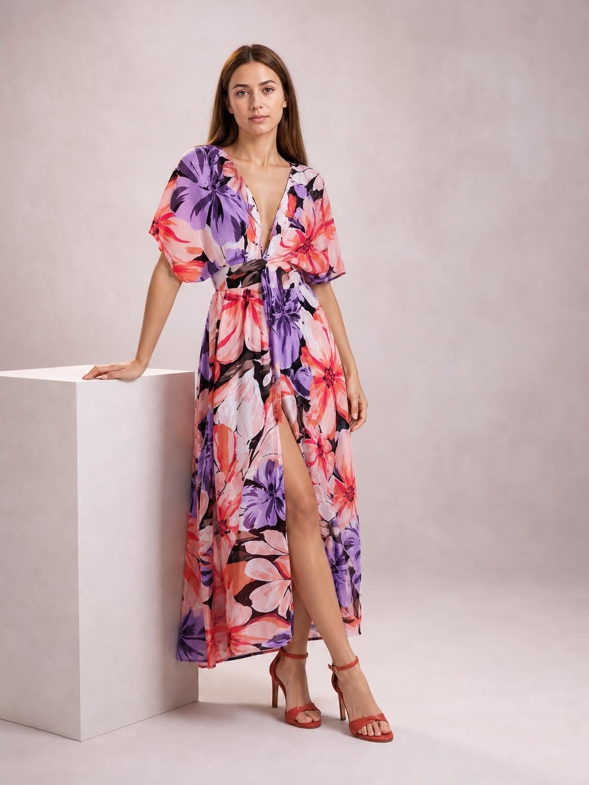 Model poses beside a pedestal in a colorful floral print maxi dress with a V-neck, twisted waist, short sleeves, flowing silhouette, and high front slit.