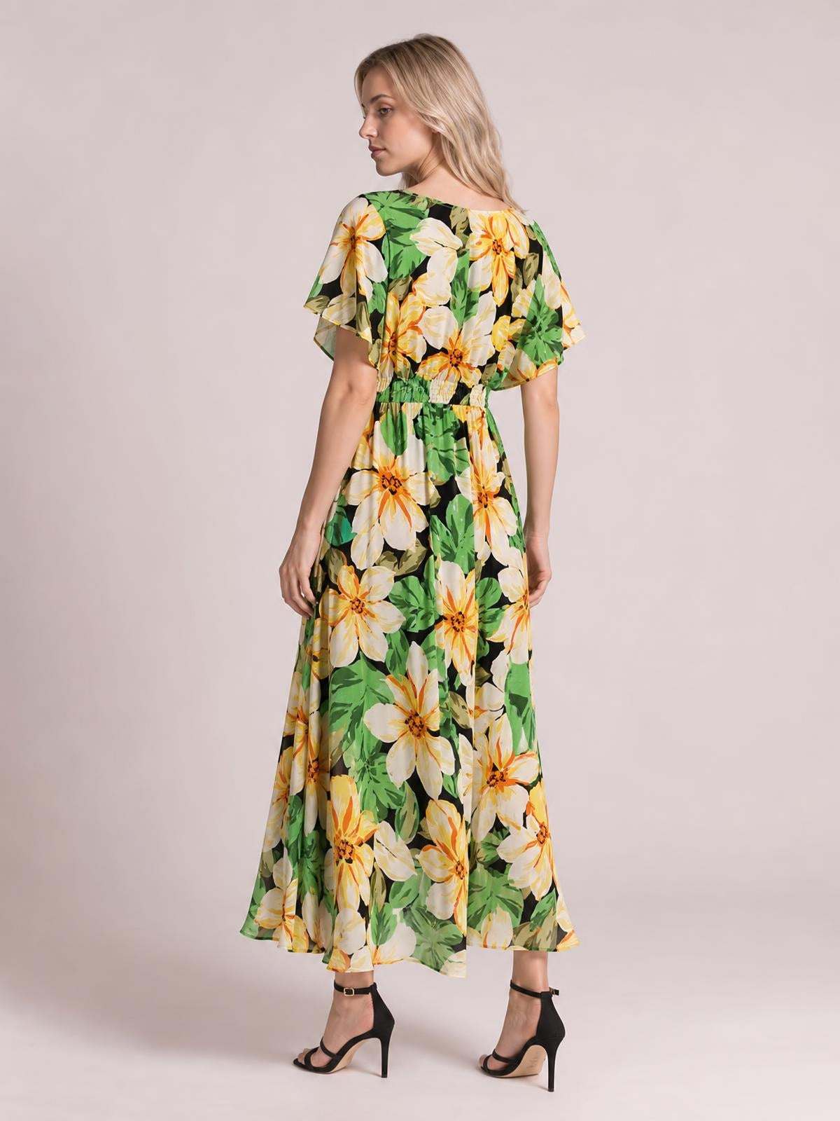 Model in a green, black, yellow, and white floral print maxi dress with flutter sleeves, a smocked waist, and a flowing ankle-length silhouette.