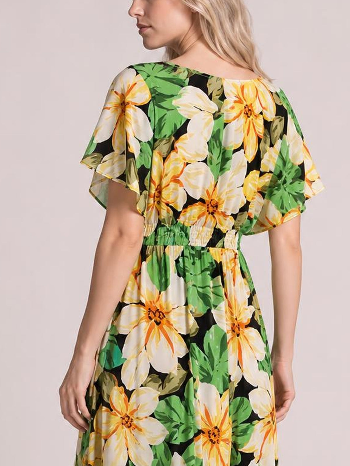Model shown from behind in a black floral print maxi dress with green leaves, cream and orange flowers, flutter sleeves, and a smocked waist creating a flowing silhouette.