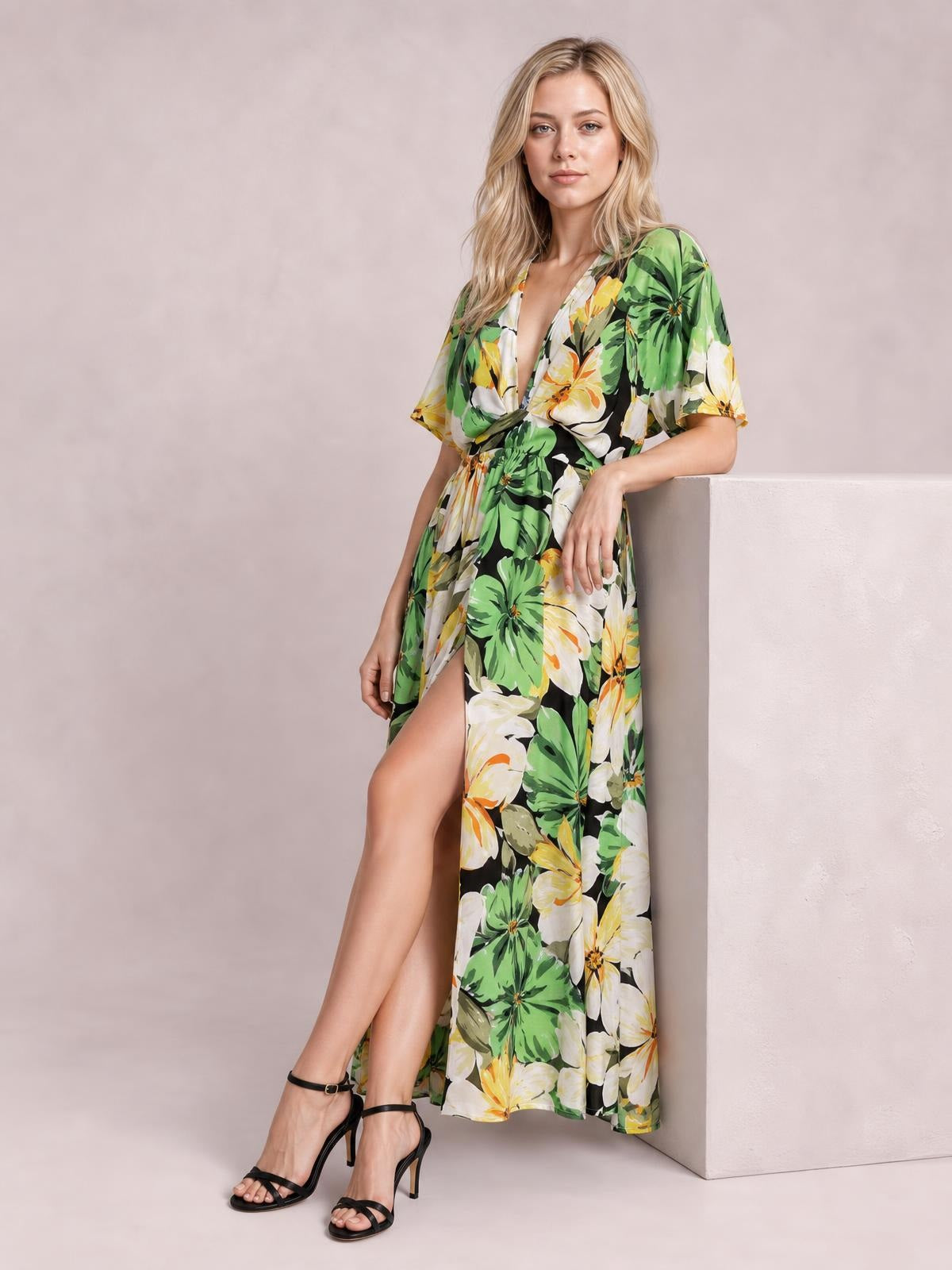 A model leans against a ledge wearing a green, black, white, and yellow floral-print V-neck maxi dress with flutter sleeves, a twisted waist, and a high side slit.