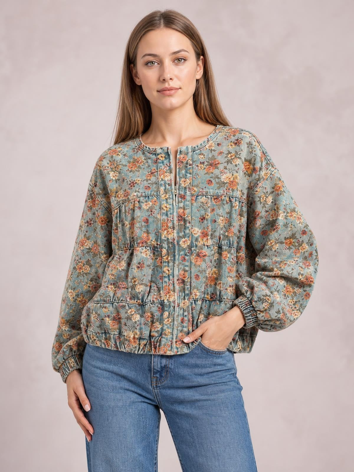 Model poses in a blue floral-print zip-up cropped denim jacket with long sleeves, elastic cuffs and hem, a round neckline, and hands in the pockets.