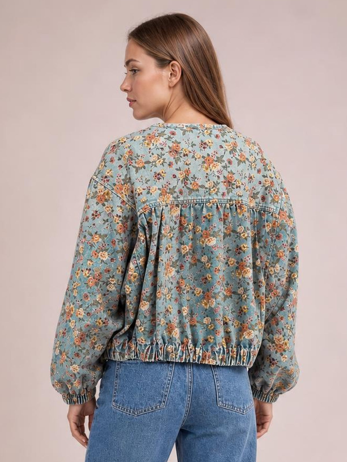 Model turns to show the back of a light blue floral print cropped denim jacket with long sleeves, gathered seams, elastic cuffs, and a zip front.