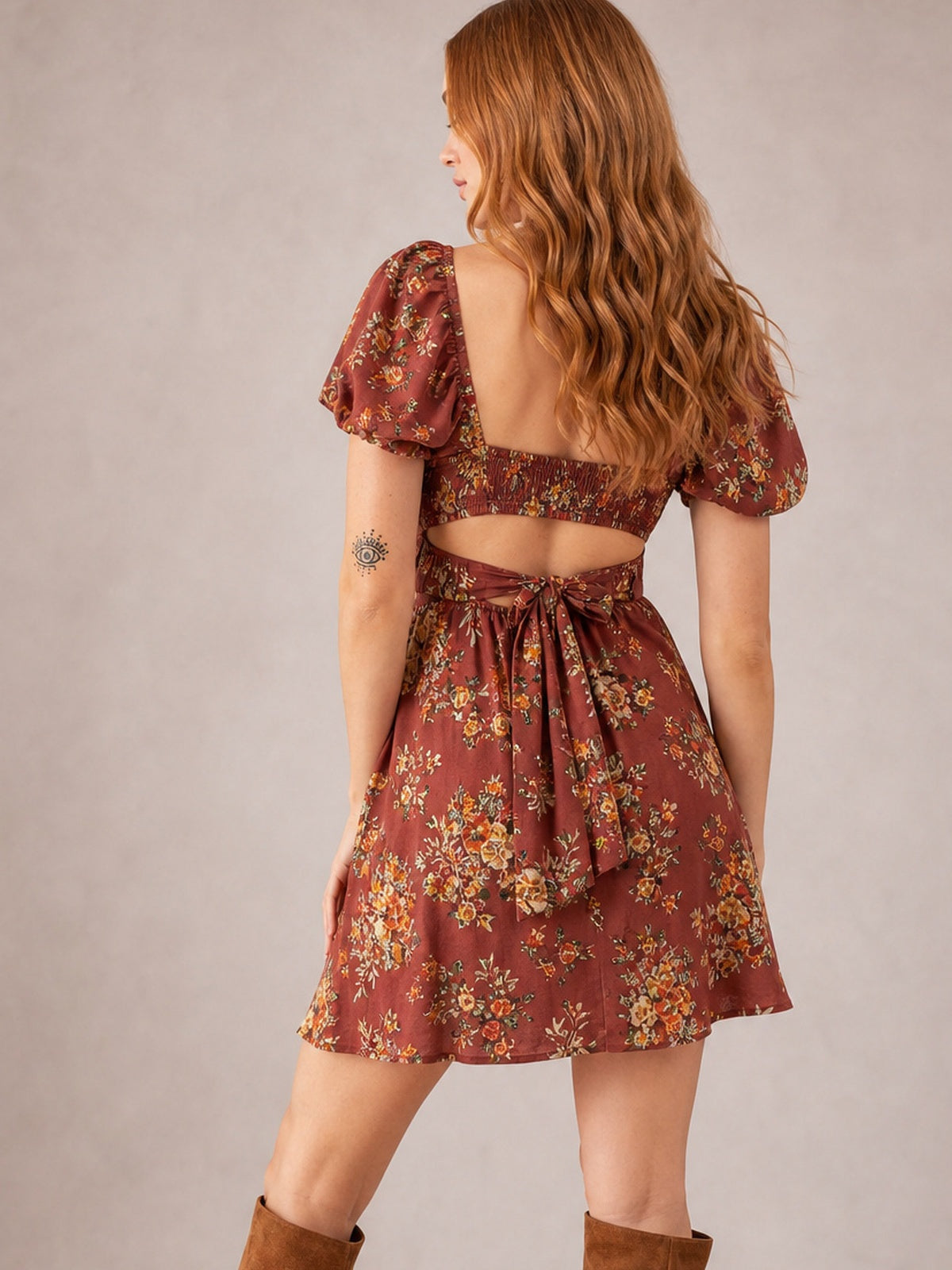 A model poses from behind in a rust floral mini dress with puff sleeves, an open back, smocked bodice, and a playful waist tie creating a flared silhouette.