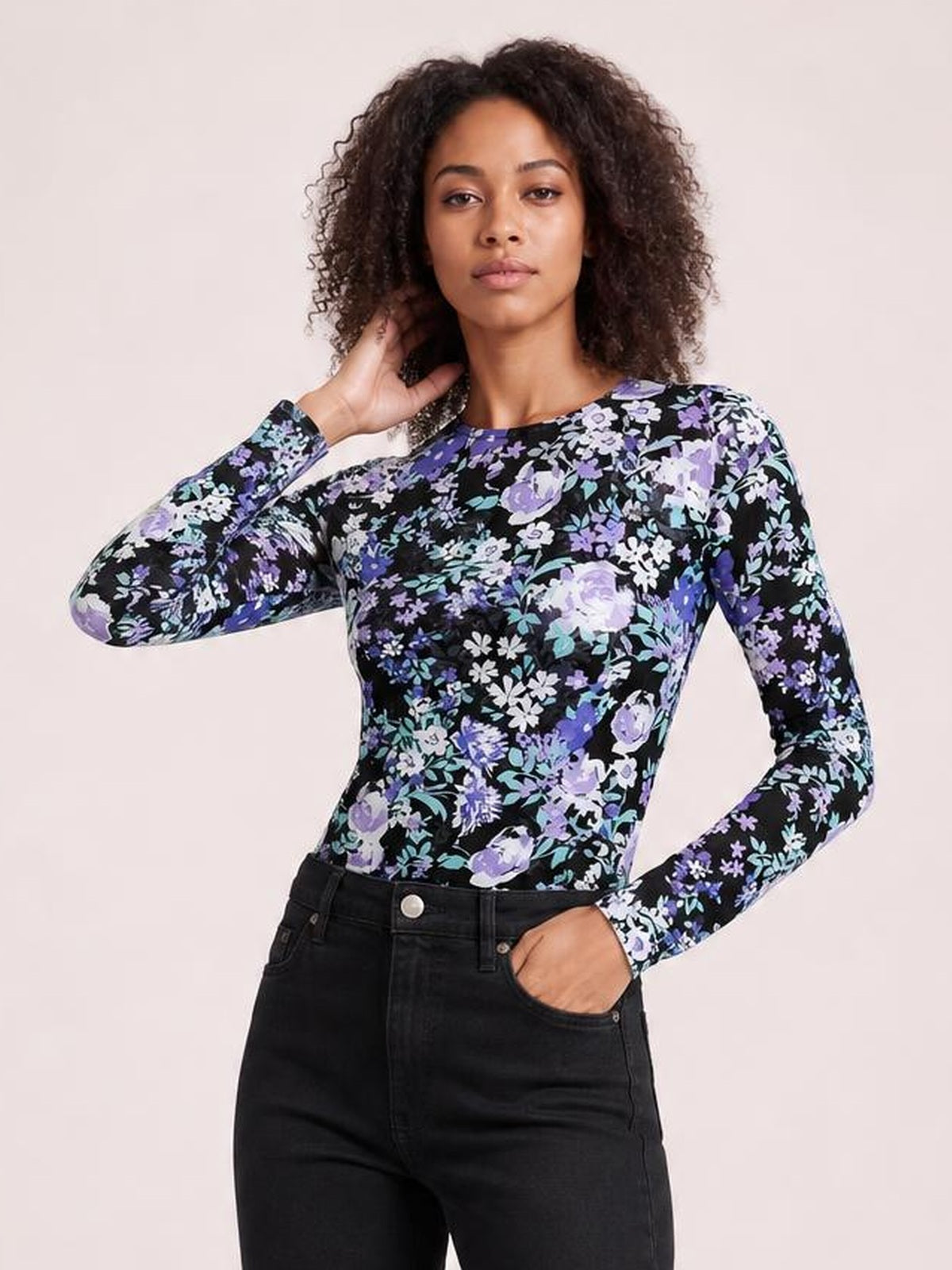 A model wears a fitted black floral round-neck long-sleeve T-shirt with purple, lavender, and blue flowers, posing with one hand in her pocket.