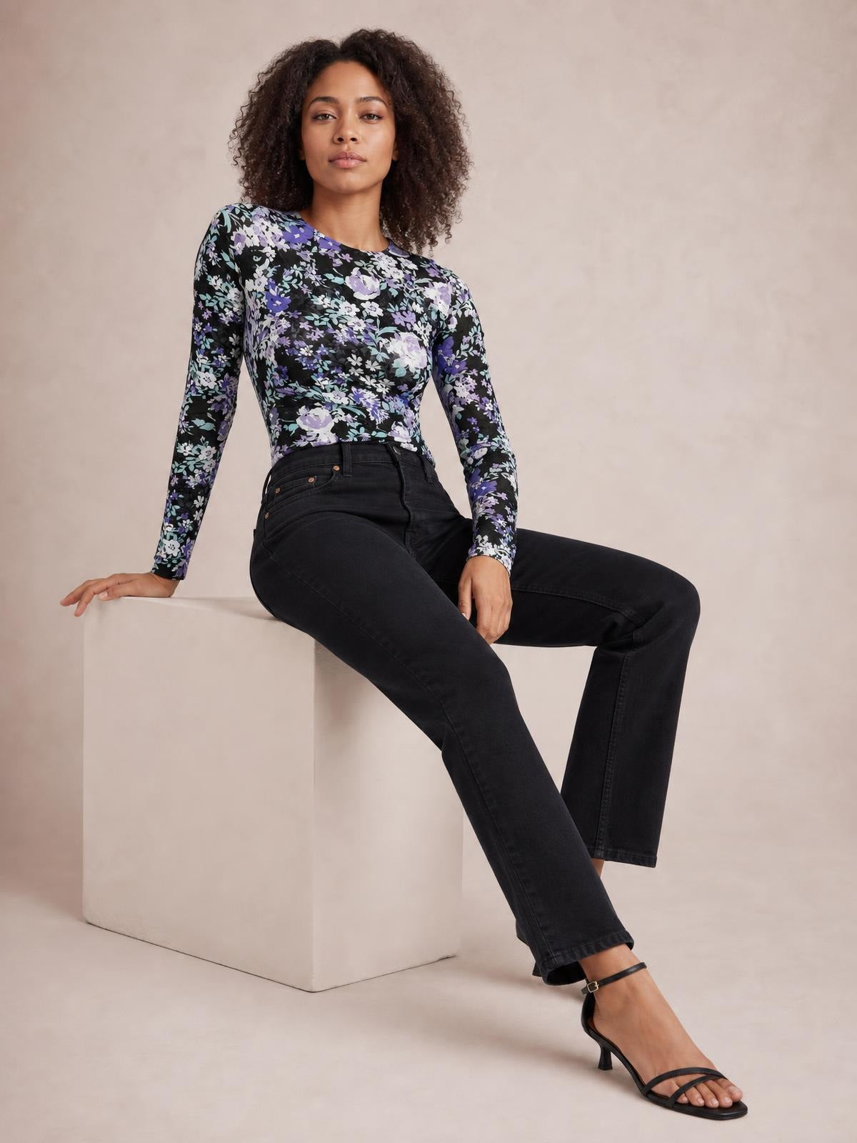 A model sits in a relaxed pose wearing a fitted black floral round neck long sleeve T-shirt with purple, white, and green blossoms.