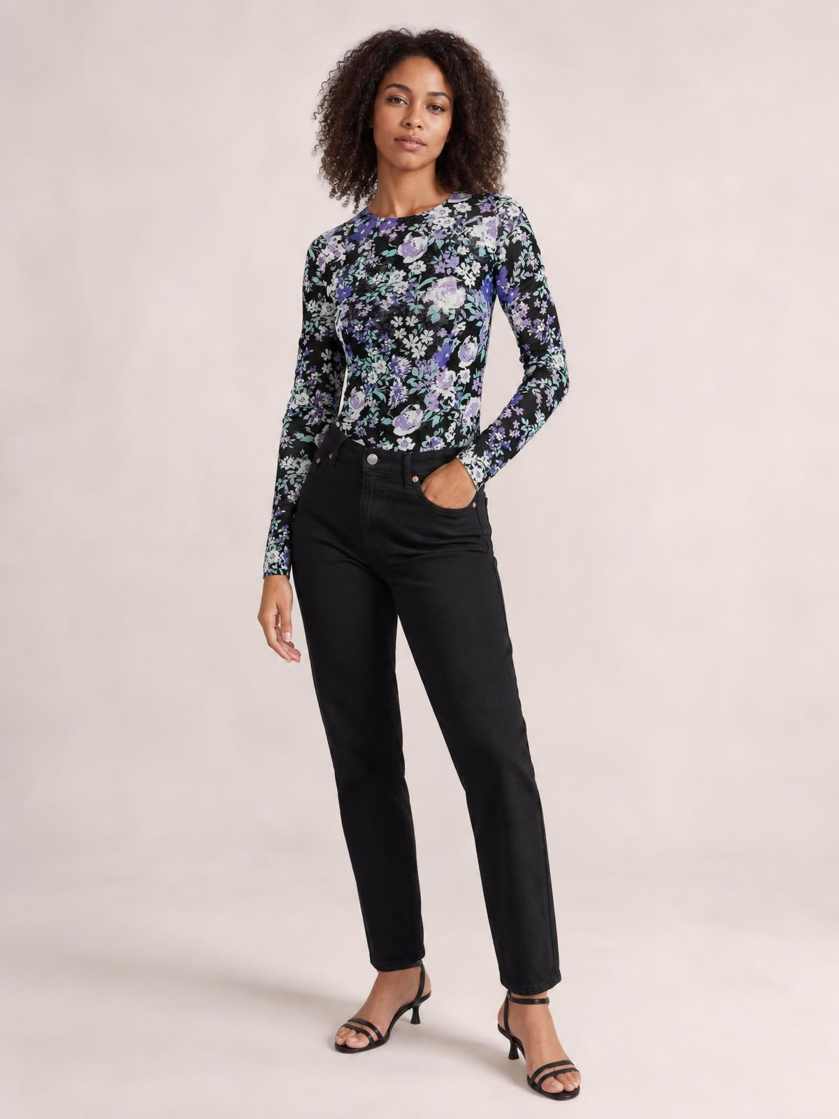 Model poses in a floral black, purple, and white round neck long sleeve T-shirt with a fitted silhouette, tucked into high-rise black pants.