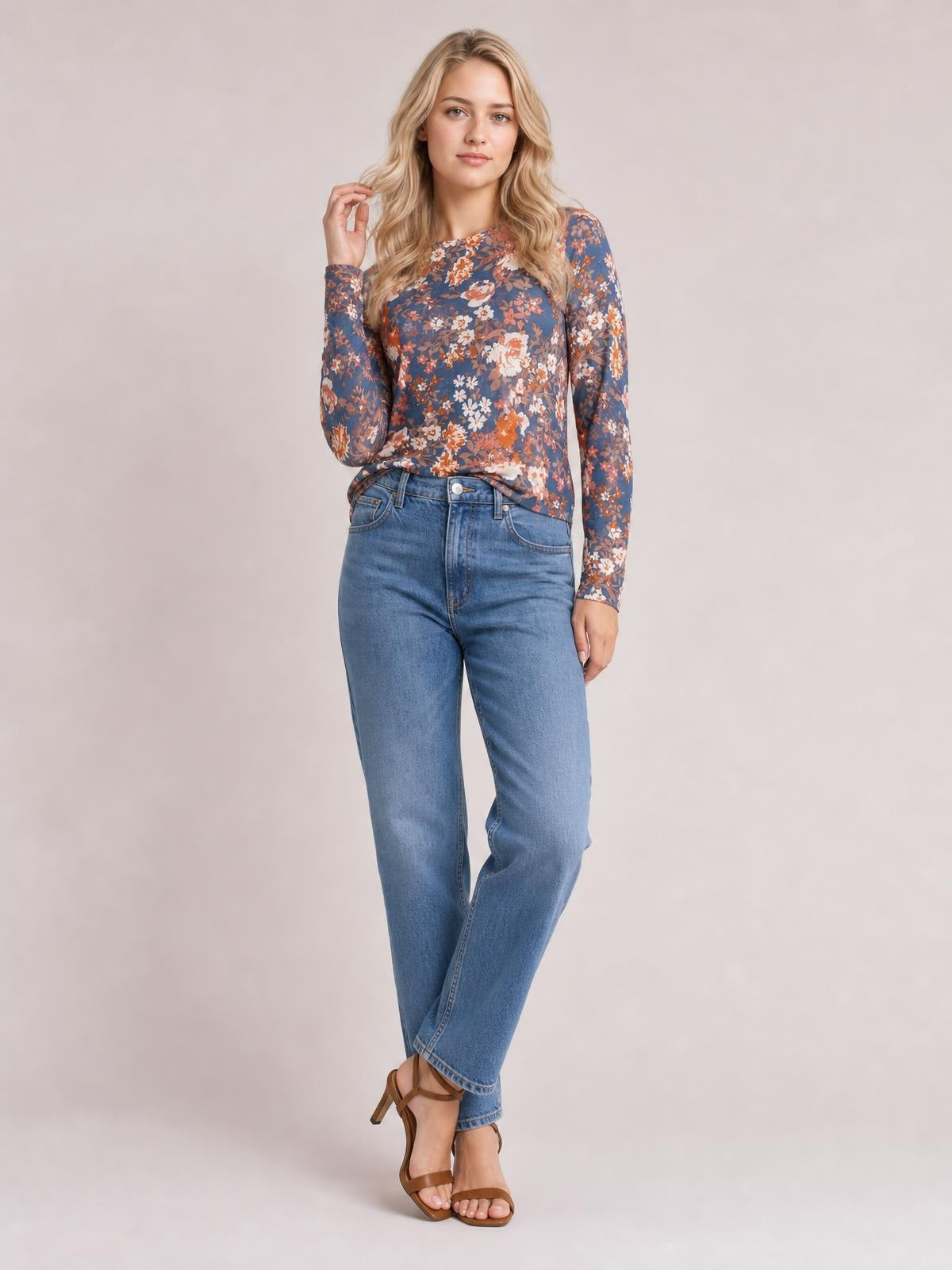 A model poses in a blue floral round-neck long-sleeve T-shirt with a fitted silhouette, gently touching her hair as she stands with crossed legs.