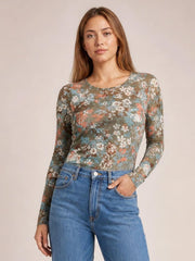 A model wears a fitted floral round neck long sleeve T-shirt in muted olive with multicolor flowers, standing front-facing with one hand in her jeans pocket.