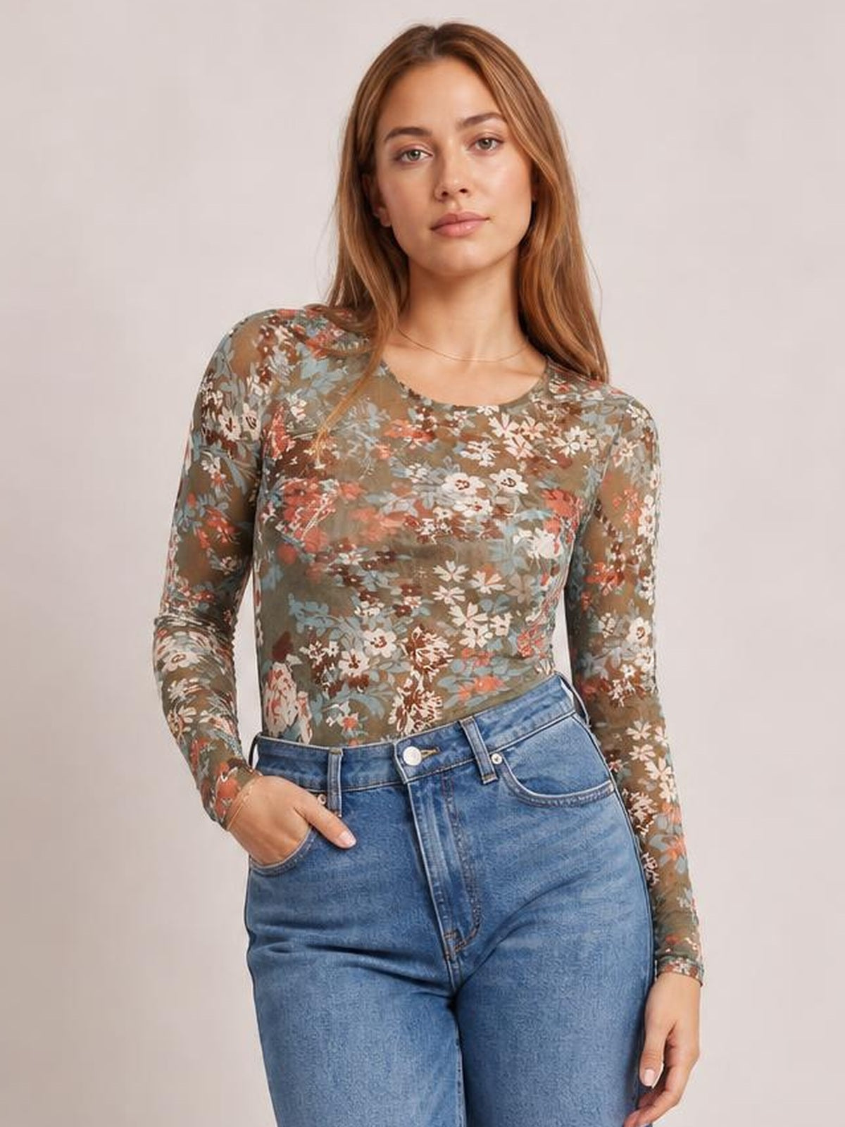 A model poses with one hand in her jeans pocket, wearing a fitted floral round neck long sleeve T-shirt in olive brown with multicolor blooms.