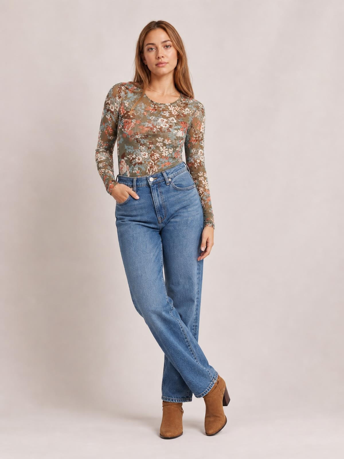 A model poses with one hand in her pocket wearing a fitted floral round-neck long-sleeve T-shirt in muted brown, olive, blue, and rust tones.