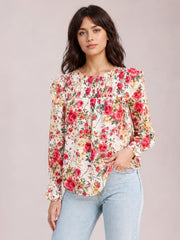 Model wearing an opaque floral round-neck smocked blouse with long sleeves, a relaxed silhouette, and a curved hem, posing with one hand in her jeans pocket.