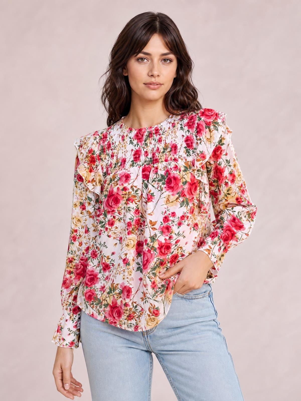 Model wears a loose floral round-neck smocked blouse in pink, white, and red with long sleeves and ruffled shoulder details, standing with one hand in her jeans pocket.