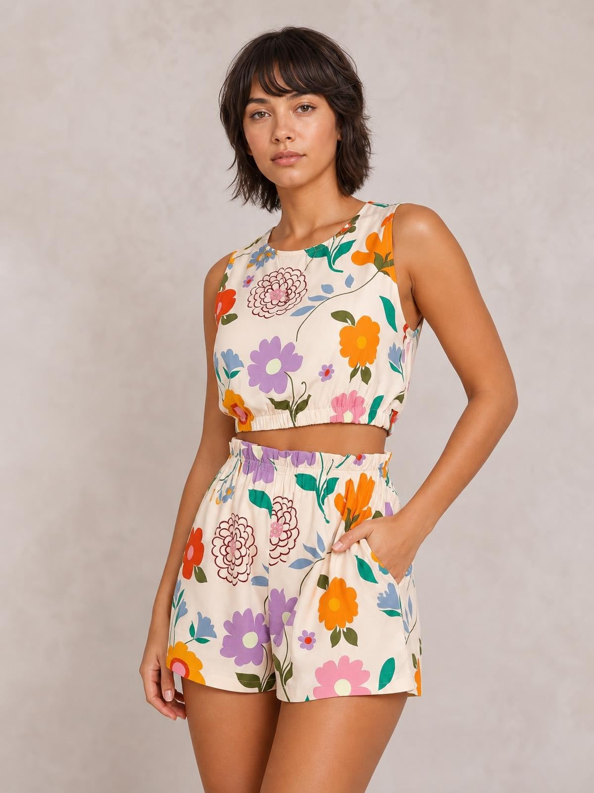 Model poses in a floral round neck tank and shorts set with a cropped ruched tank, colorful flower print, elastic waist, and side pockets.