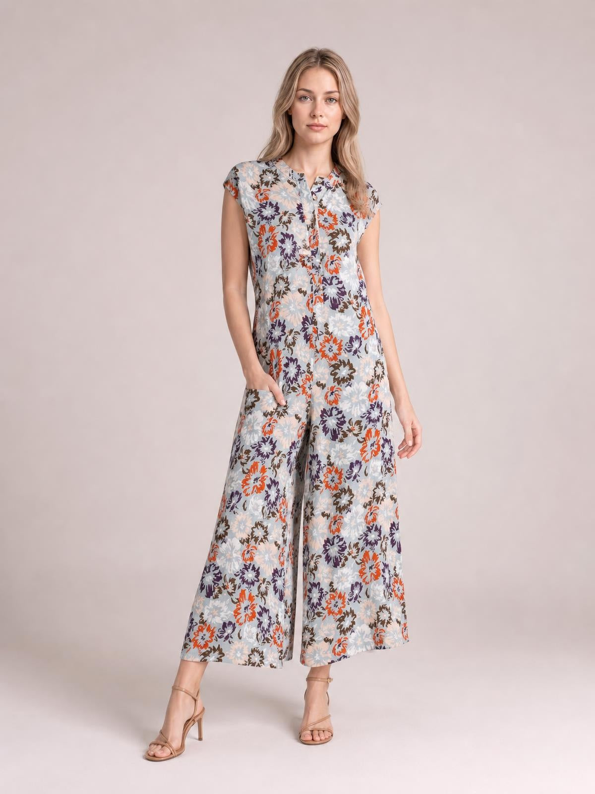 Model wears a light blue floral sleeveless jumpsuit with a button front, softly tailored bodice, and cropped wide-leg silhouette, posing with one hand at her hip.