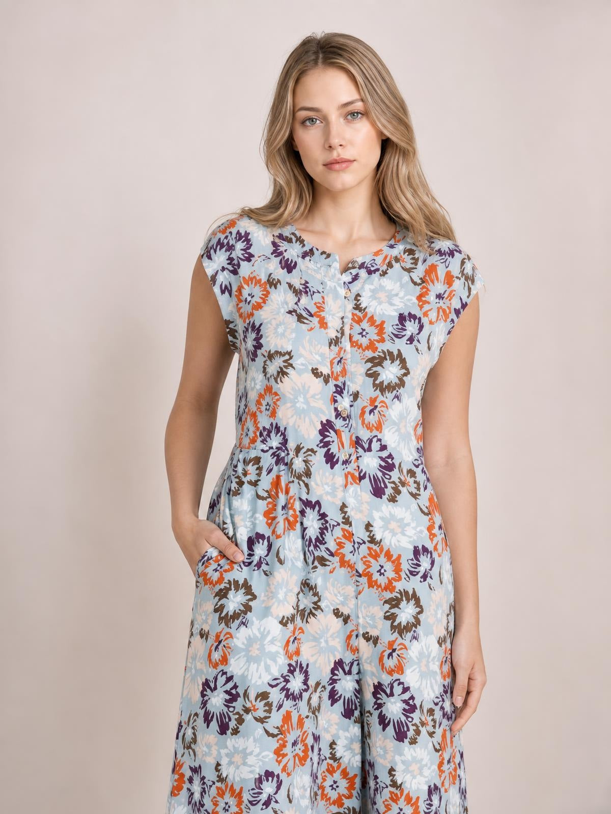 Model wears a light blue floral sleeveless button-front jumpsuit with a fitted waist, opaque fabric, and wide legs, posing with one hand in a pocket.