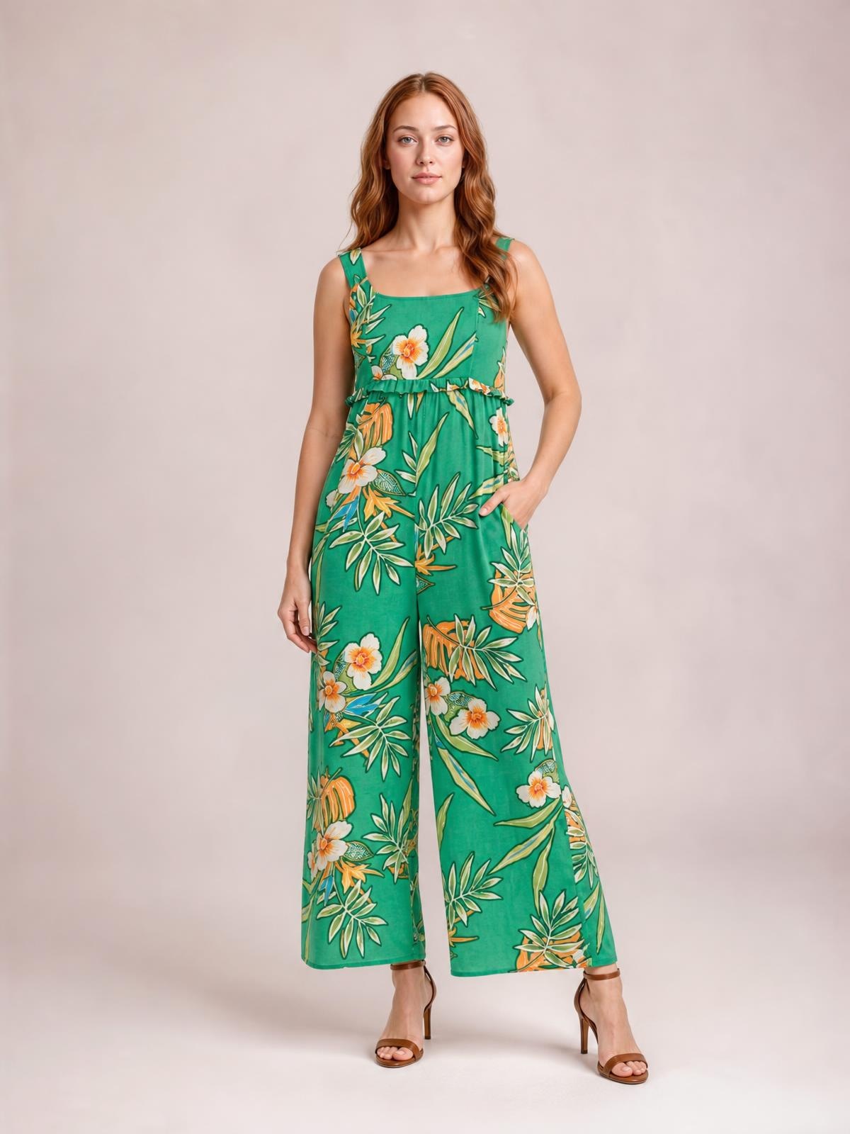 Model poses in a green floral sleeveless jumpsuit with a ruffled waist, pockets, square neckline, and relaxed wide-leg silhouette.