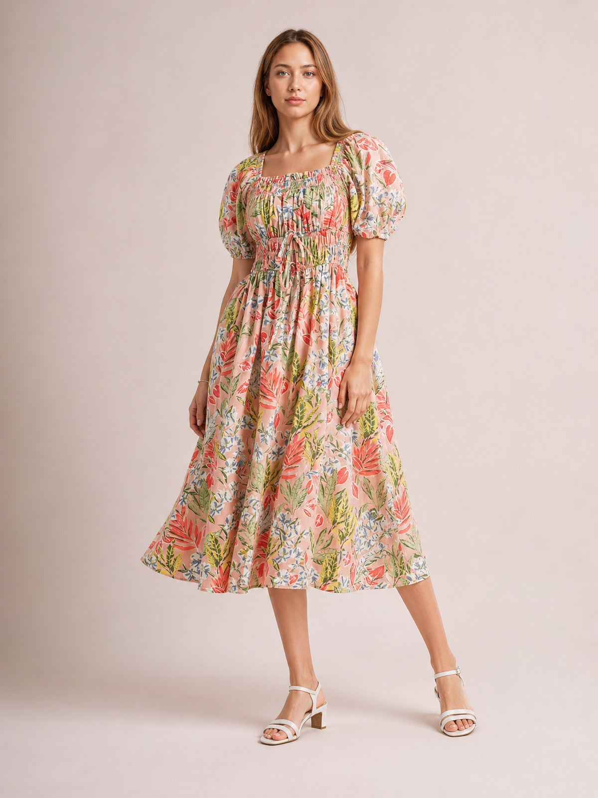 Model poses in a floral smocked midi dress with a square neckline, puff short sleeves, tied waist, and flowing coral, pink, green, and cream skirt.