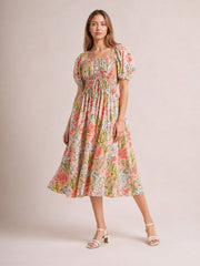 Model poses in a floral smocked midi dress with a square neckline, puff short sleeves, tied waist, and flowing coral, pink, green, and cream skirt.