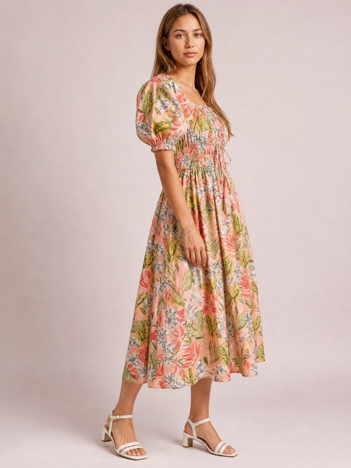 Model poses in a floral smocked midi dress with a square neckline, puff short sleeves, tied waist, and flowing gathered skirt in coral, pink, green, and cream.