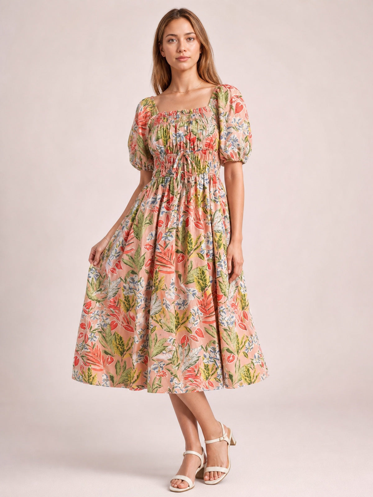 Model poses in a floral smocked midi dress with a square neckline, puff short sleeves, tied waist, and flowing gathered skirt in coral, pink, green, and cream.