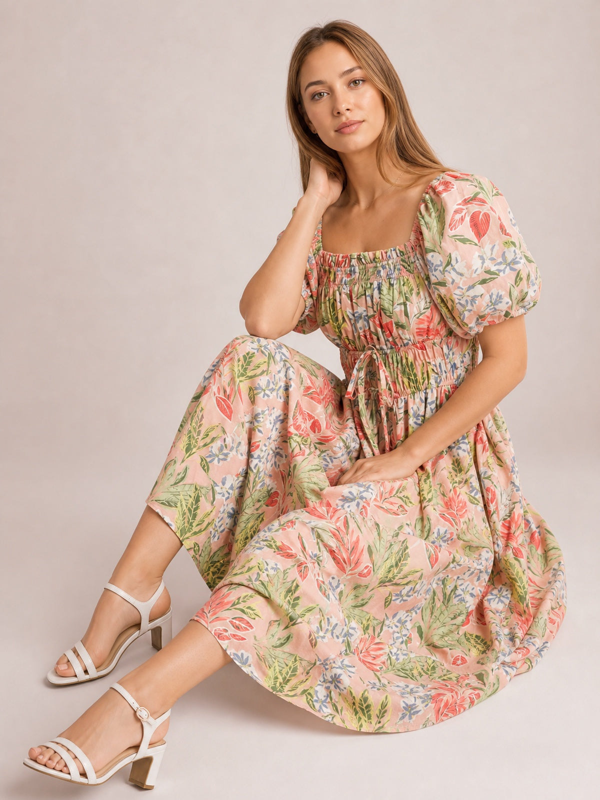 A seated model wears a coral, pink, green, and cream floral smocked midi dress with a square neck, puff short sleeves, tied waist, and flowing gathered skirt.