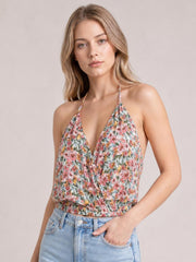 Model wears a multicolor floral surplice-neck cami with thin straps and a defined waist, paired with jeans as she poses with one hand in a pocket.