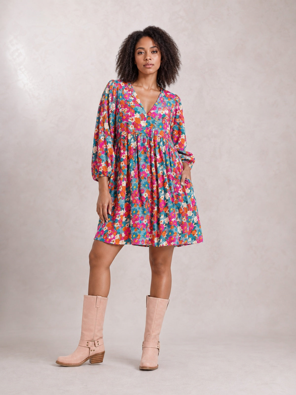A model poses in a colorful floral tie neck mini dress with a V-neck, puff sleeves, gathered waist, and flared silhouette in teal, pink, orange, and white.