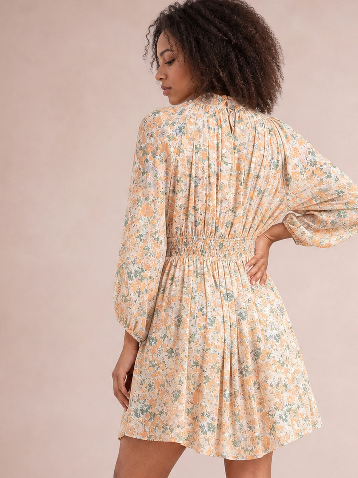 A model poses in a peach floral mini dress with voluminous balloon sleeves, a ruched waist, and a gently flared silhouette, shown from the back.
