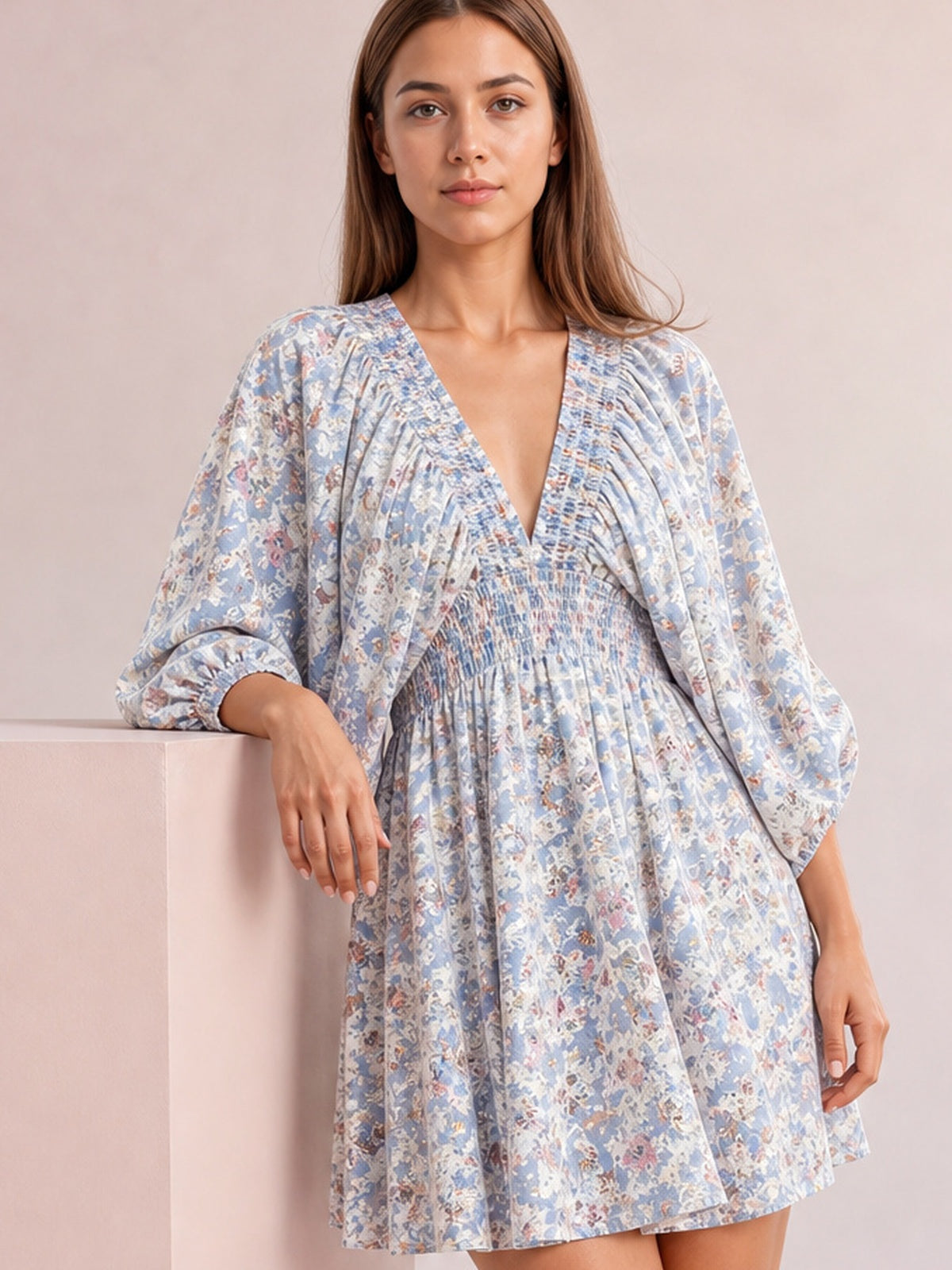 Model wearing a blue floral V-neck mini dress with voluminous balloon sleeves, ruched waist shaping, and a softly flared silhouette while leaning on a pedestal.