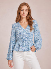 Model poses in a blue and white floral V-neck peplum blouse with long balloon sleeves, gathered waist, flared hem, and one hand in her pocket.