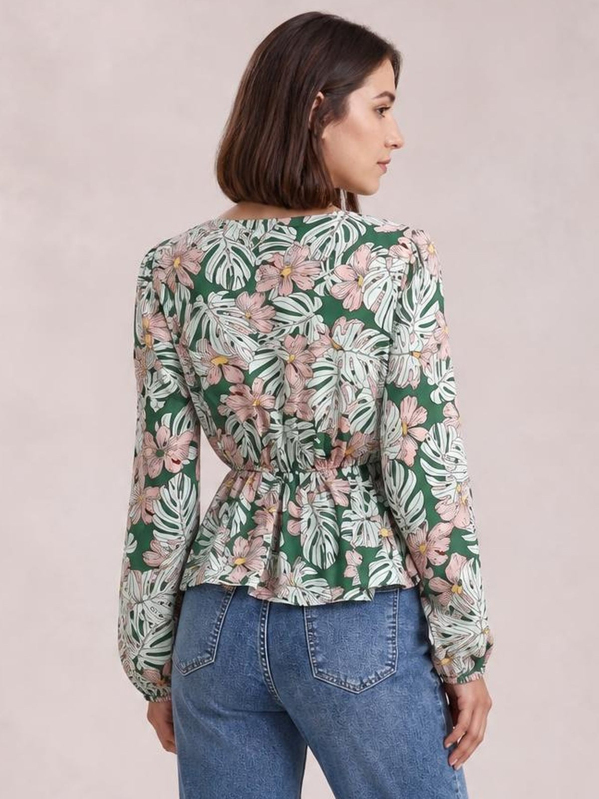 Model shown from behind wearing a green floral peplum blouse with long balloon sleeves, a gathered waist, and a flared hem over blue jeans.