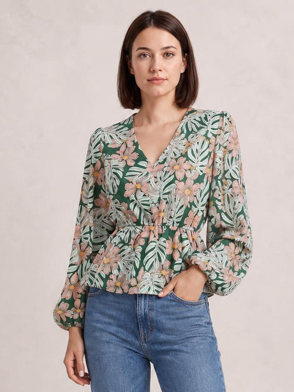 Model wearing a green floral V-neck peplum blouse with voluminous balloon sleeves and a gently flared silhouette, standing with one hand in her jeans pocket.
