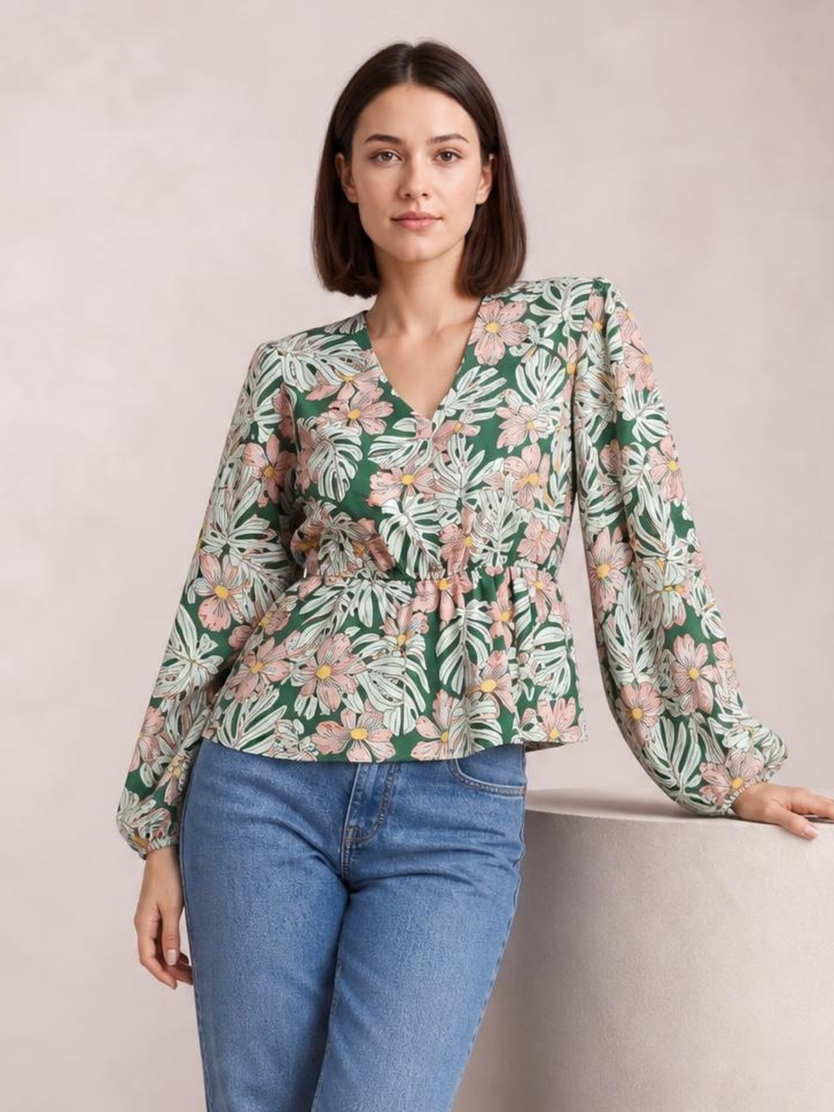 Model wears a green floral V-neck peplum blouse with voluminous balloon sleeves and gathered cuffs, standing with one hand resting beside her.