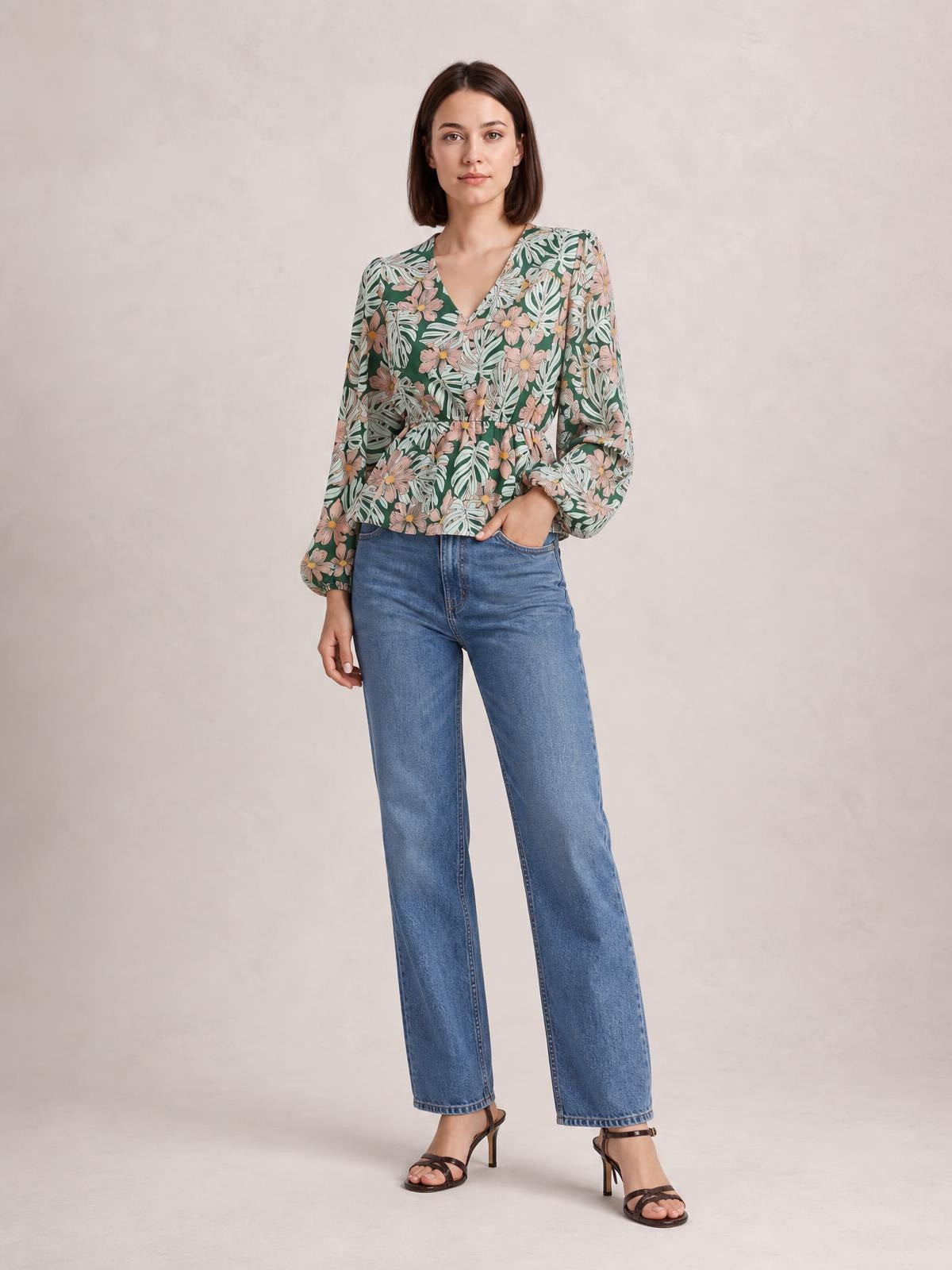 Model wearing a green floral V-neck peplum blouse with voluminous balloon sleeves, gathered waist, and a softly flared silhouette while standing with one hand in her pocket.