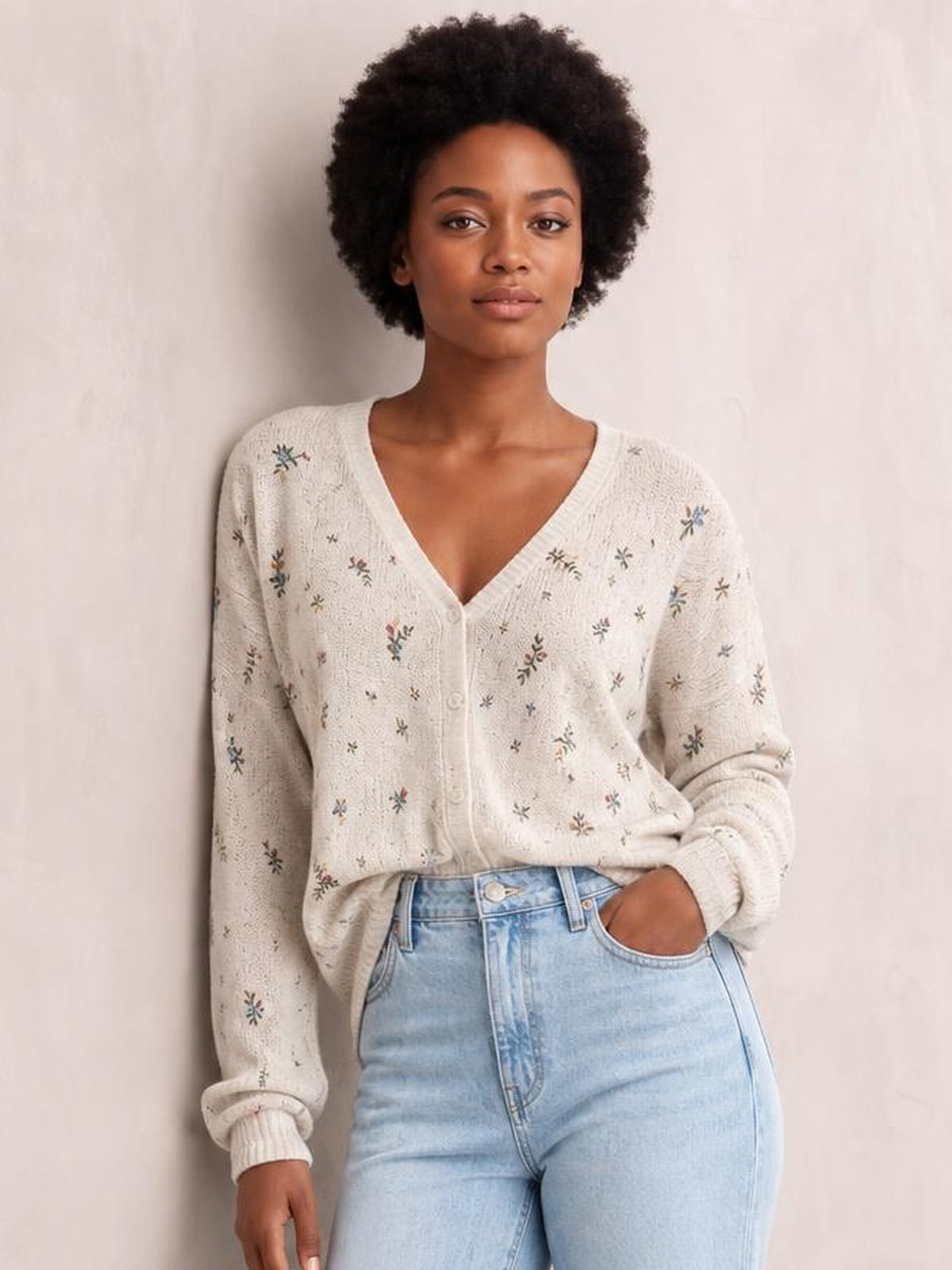 Model poses with one hand in her jeans pocket, wearing a cream floral V-neck button-down cardigan with long sleeves, ribbed cuffs, and a relaxed silhouette.