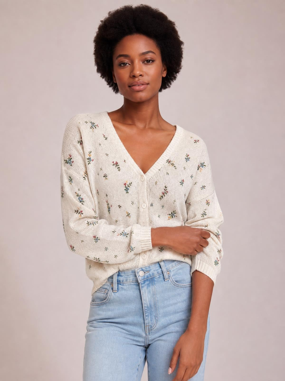 A model poses with folded arms in an ivory floral V-neck cardigan featuring button-down construction, long sleeves, ribbed edges, and a relaxed silhouette.